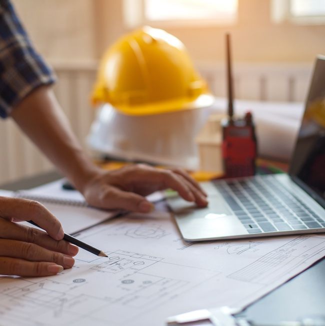 A person is sitting at a table with a laptop and a hard hat in the background.