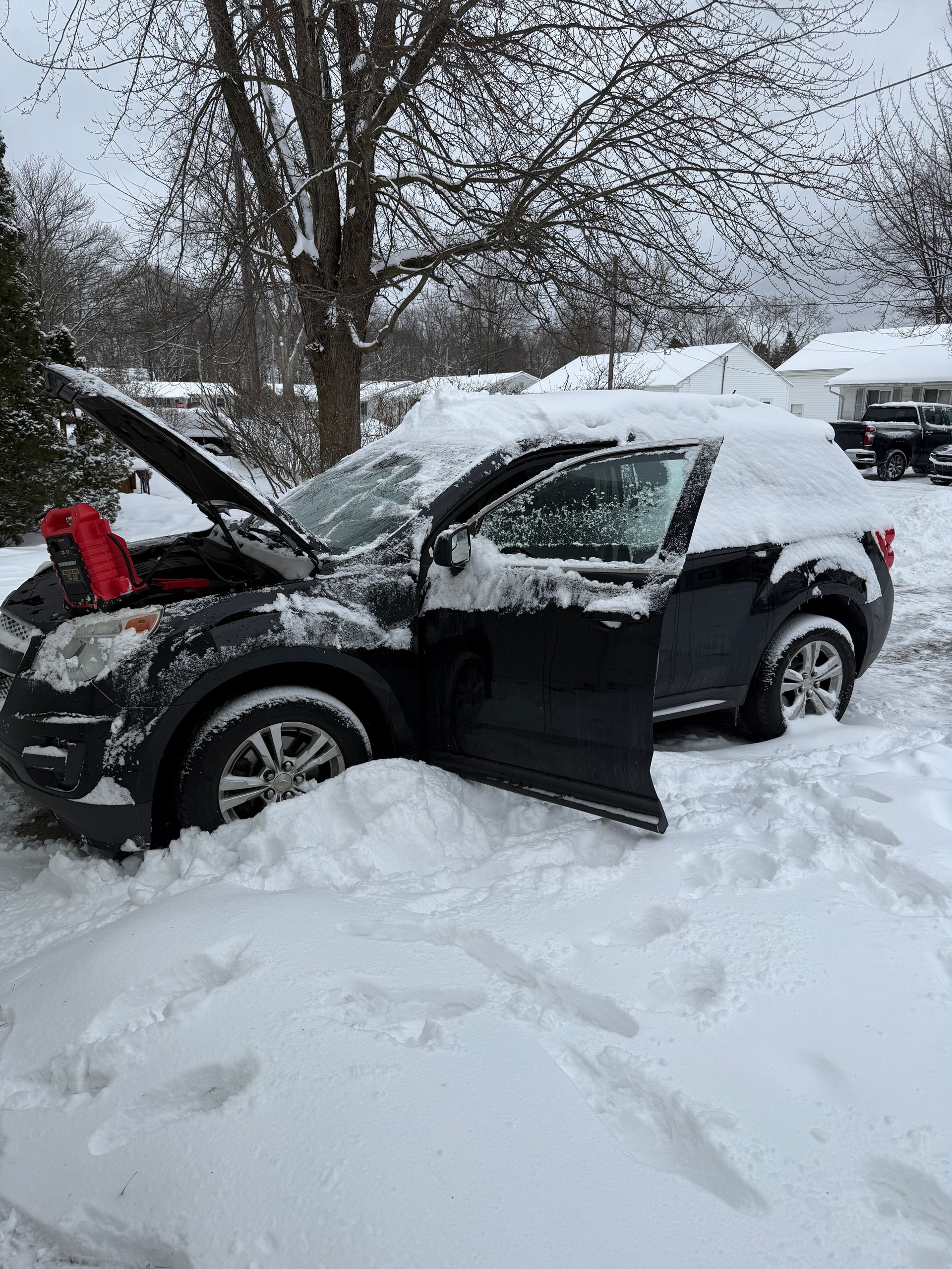 A black SUV with its hood and driver-side door open, parked in a deep, snow-covered residential area during a snowfall