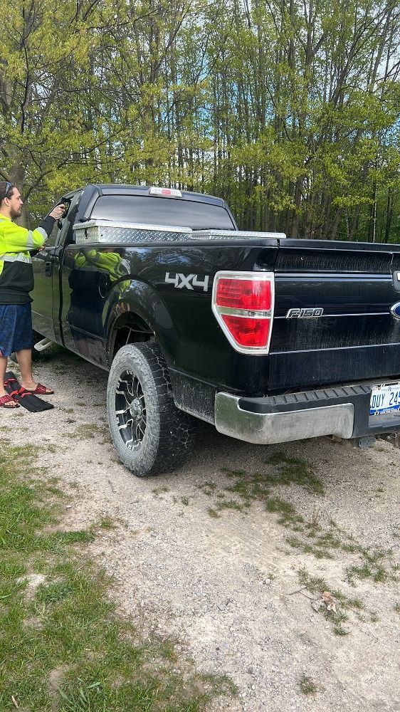 A person in a high-visibility yellow jacket stands beside a black Ford pickup truck on a gravel path near trees.