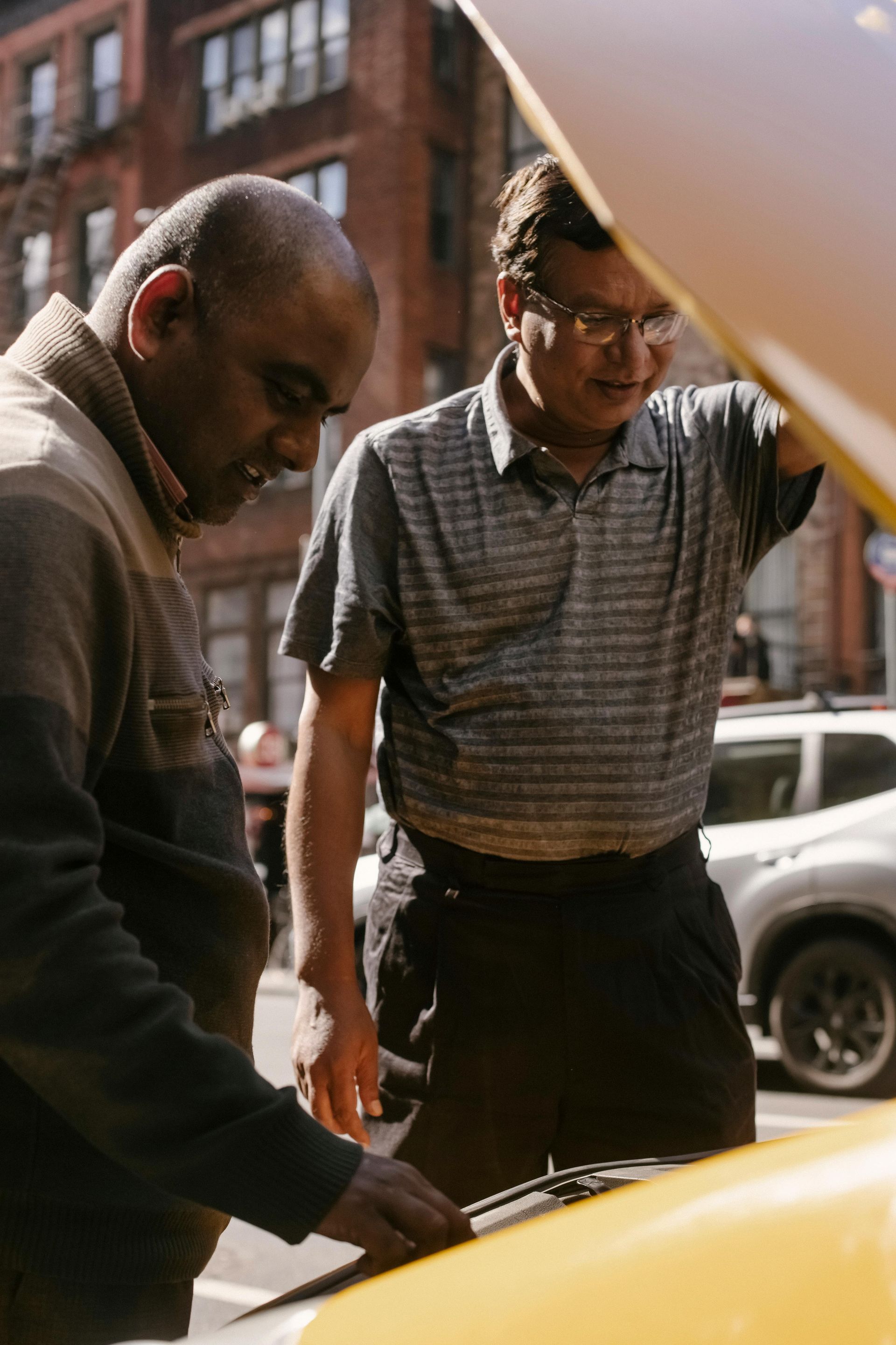 Two men are looking under the hood of a yellow car.