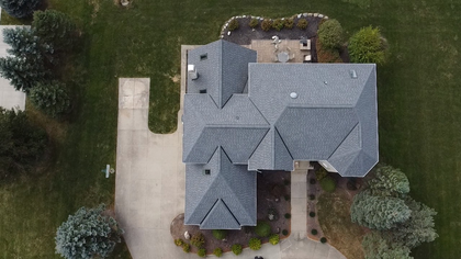 Aerial view of a gray-roofed house with driveway, front walkway, and surrounding trees.
