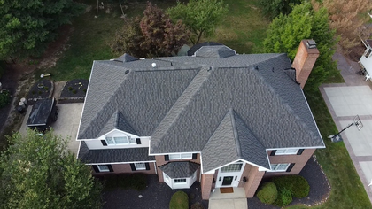 Aerial view of a gray-roofed brick house with a driveway and surrounding trees.
