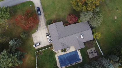 Aerial view of a house with a blue pool, driveway, lawn, and trees surrounding it