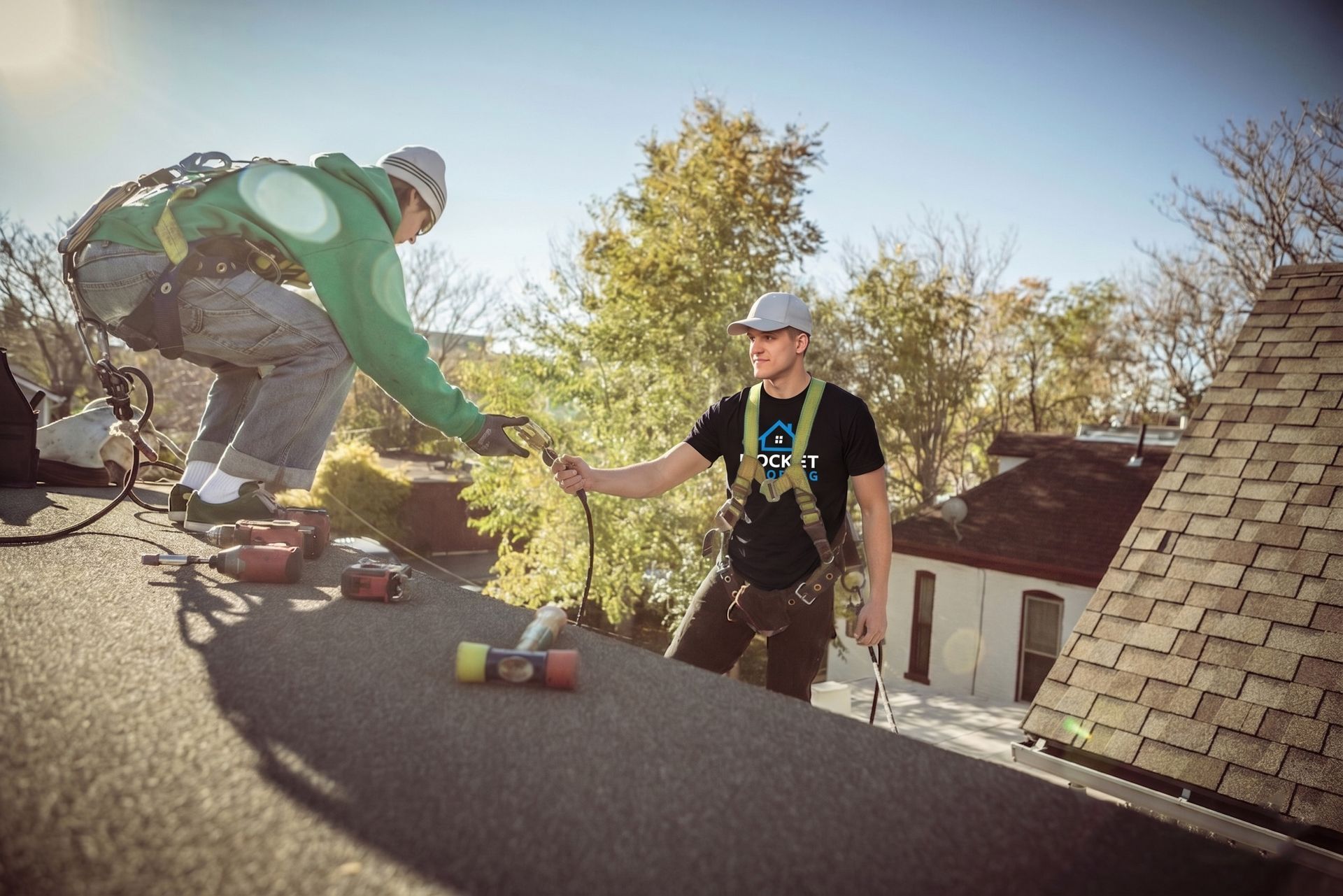 Two roofers on a rooftop passing tools, with shingles and a house in the background.