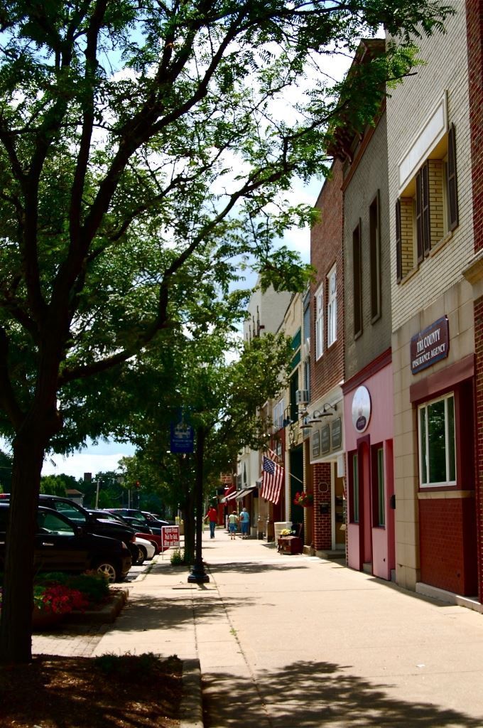 Sunlit sidewalk beside brick storefronts and parked cars under leafy trees