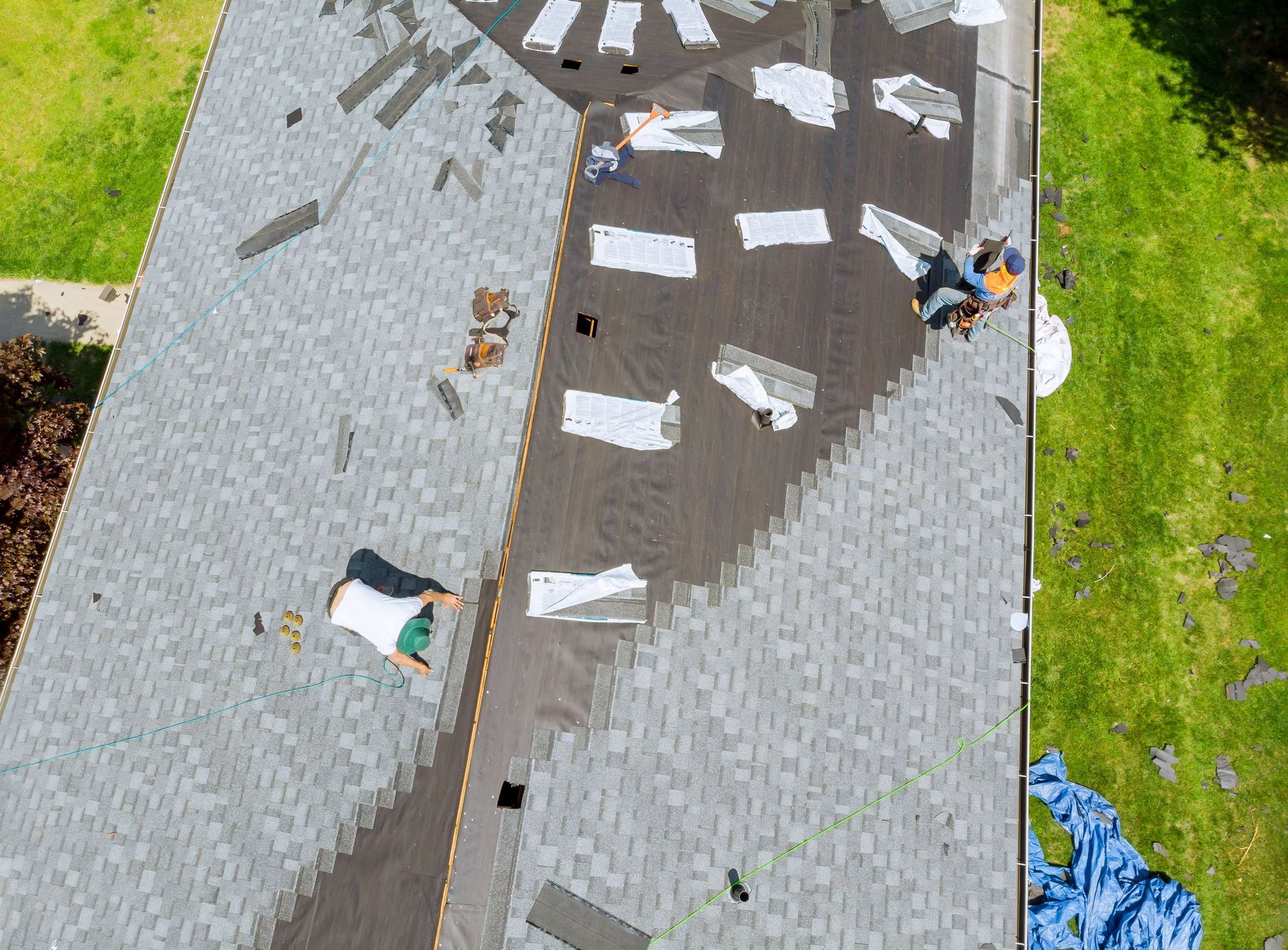 Workers installing shingles on a gray roof, with scattered white materials and green lawn below.