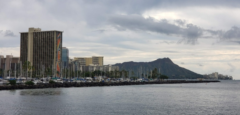 Coastal city skyline with high-rises, a mountain backdrop, and calm water under cloudy skies