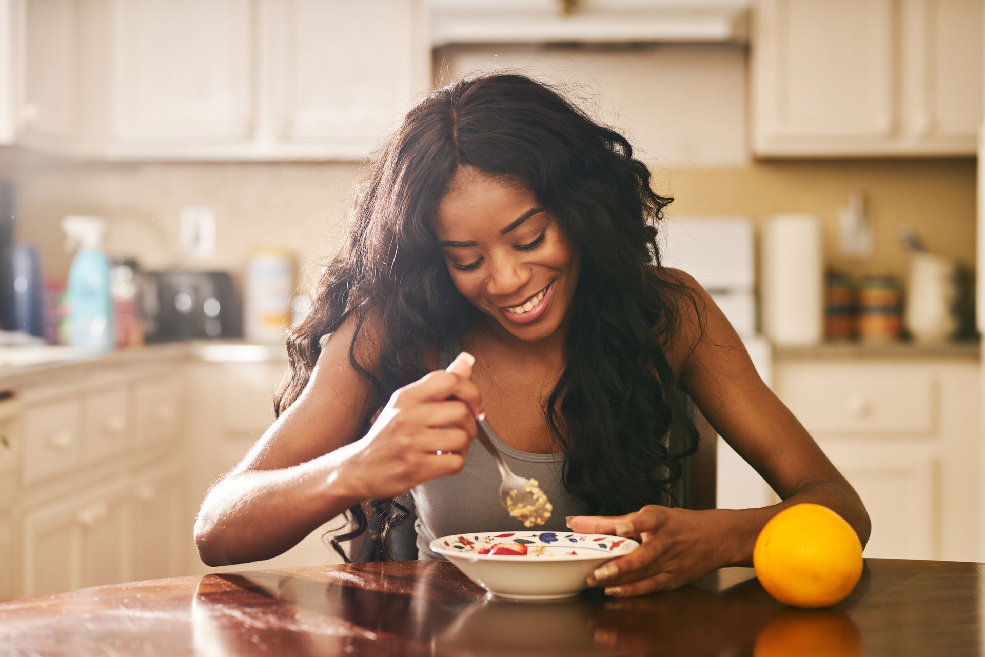 Woman in workout clothes eats cereal with fruit, smiling, next to dumbbells.