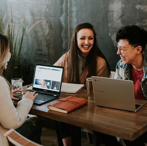 Three people laughing while working at a table with laptops, notebooks, and drinks.