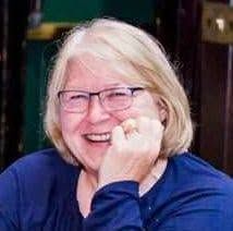 Pam smiling with blonde hair and glasses, resting chin on hand, wearing a blue top.