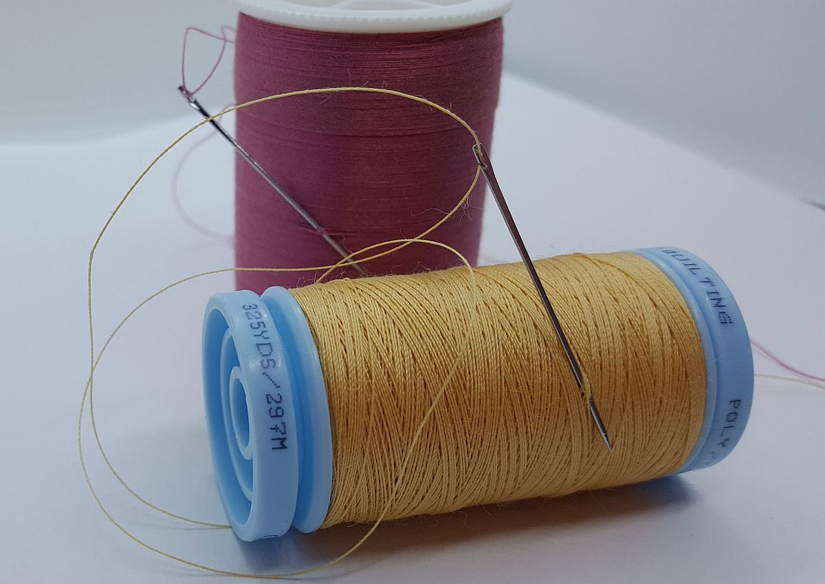 Two spools of thread, gold and burgundy, with a needle and loose thread on a white surface