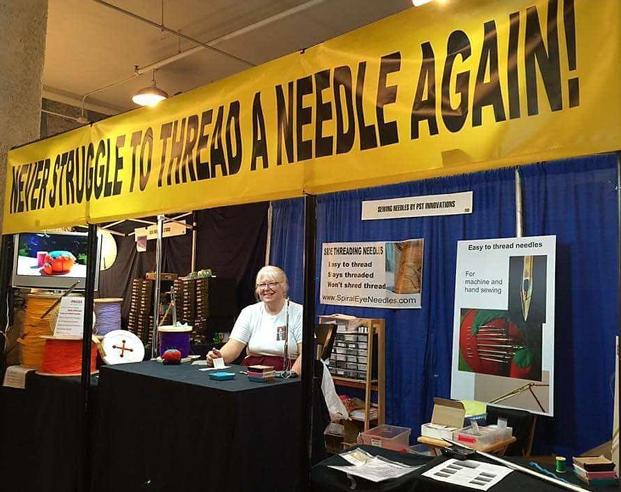 Trade show booth with a yellow “Needle again!” banner, display posters, and a seated exhibitor behind a table.