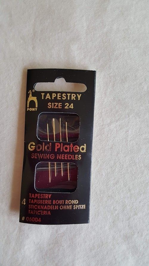 Gold-plated tapestry sewing needles package on white fabric background.