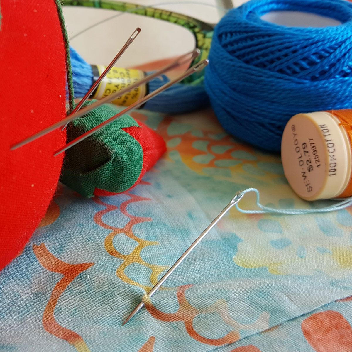Sewing supplies on patterned fabric: blue thread spool, needles, red pincushion, and green thimble