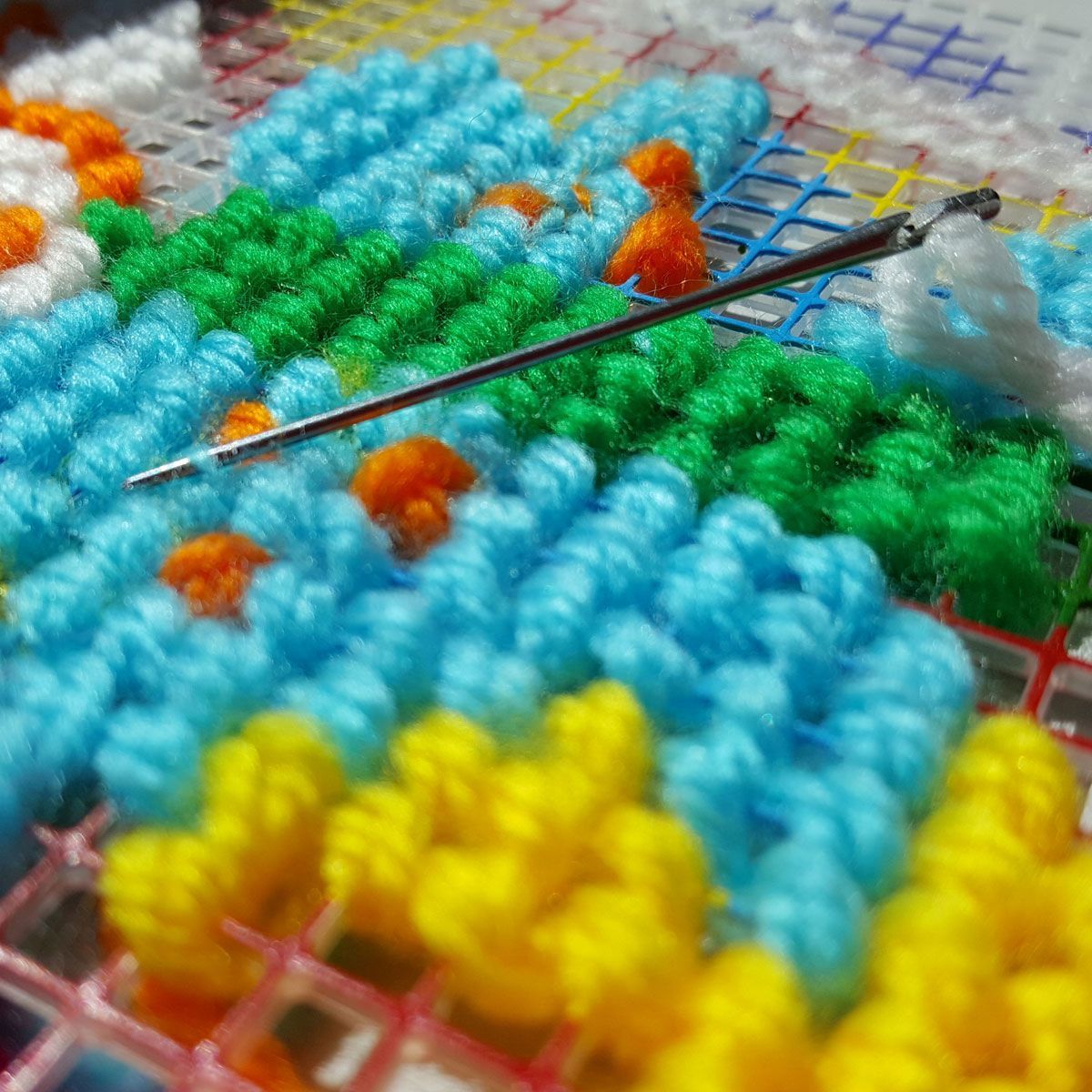 Colorful needlework canvas with a needle and thread laid across blue, green, yellow, and orange stitches