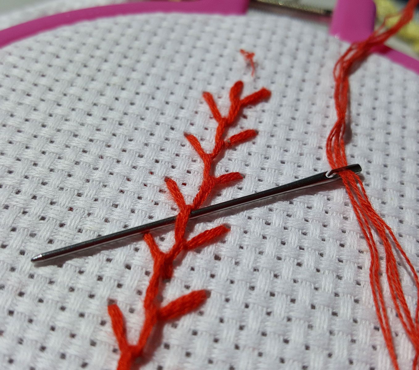 Needle and red thread stitching a leafy vine on white fabric in close-up.