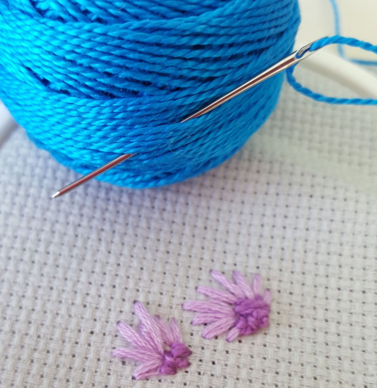 Blue thread spool with a needle, above two small pink embroidered flowers on fabric