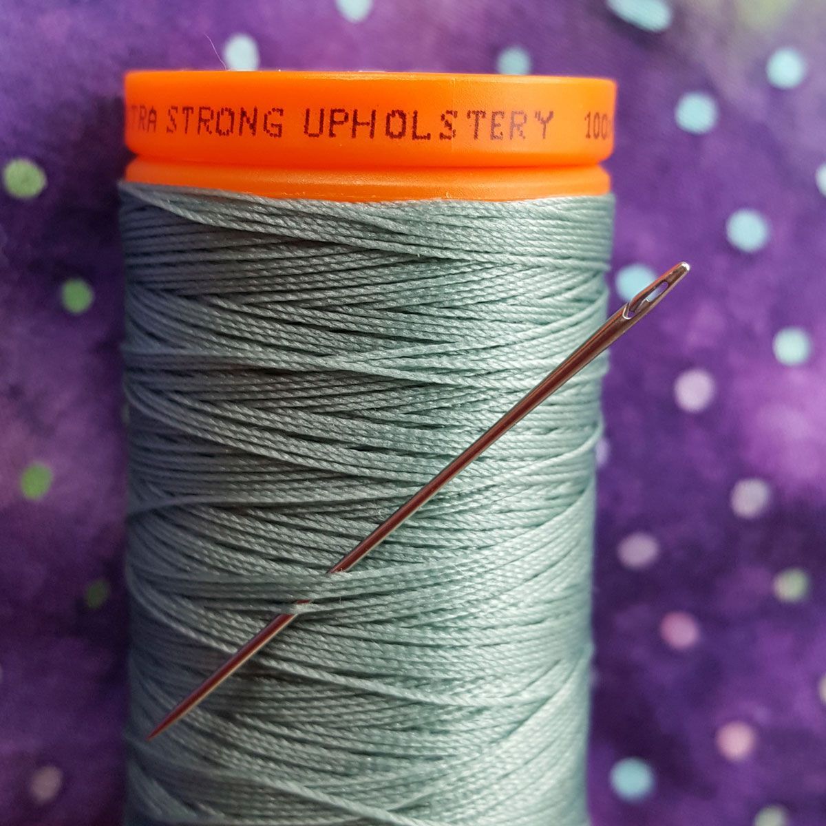 Spool of gray sewing thread with an orange top and a needle stuck through it on a purple polka-dot background