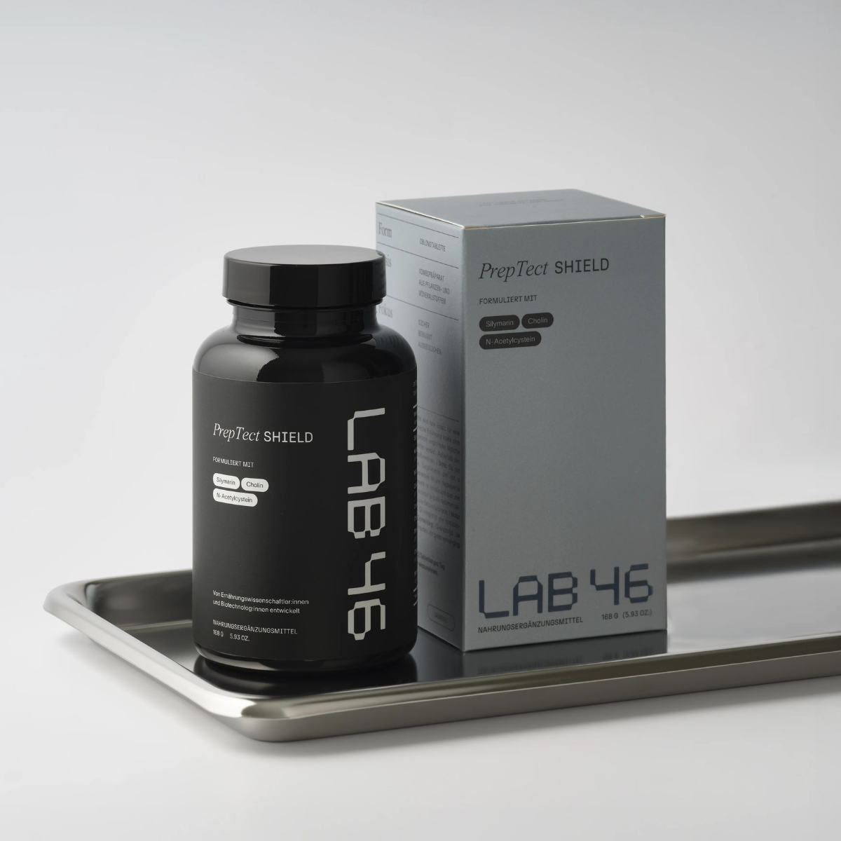 Schwarze Nahrungsergänzungsflasche und graue LAB 46-Box auf einem silbernen Tablett vor weißem Hintergrund