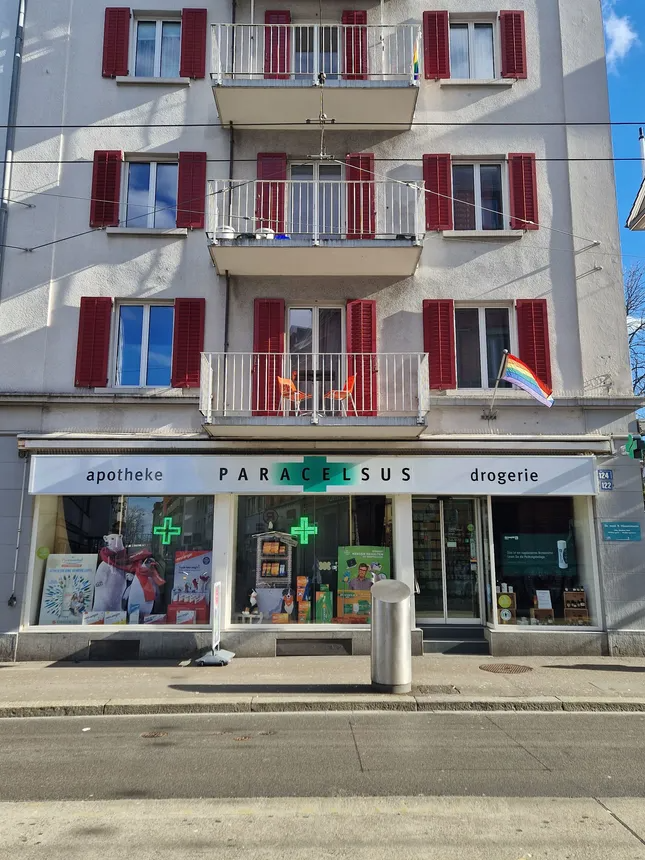 Strassenansicht einer Apothekenfassade mit darüberliegenden Balkonen und einer Regenbogenflagge im Fenster