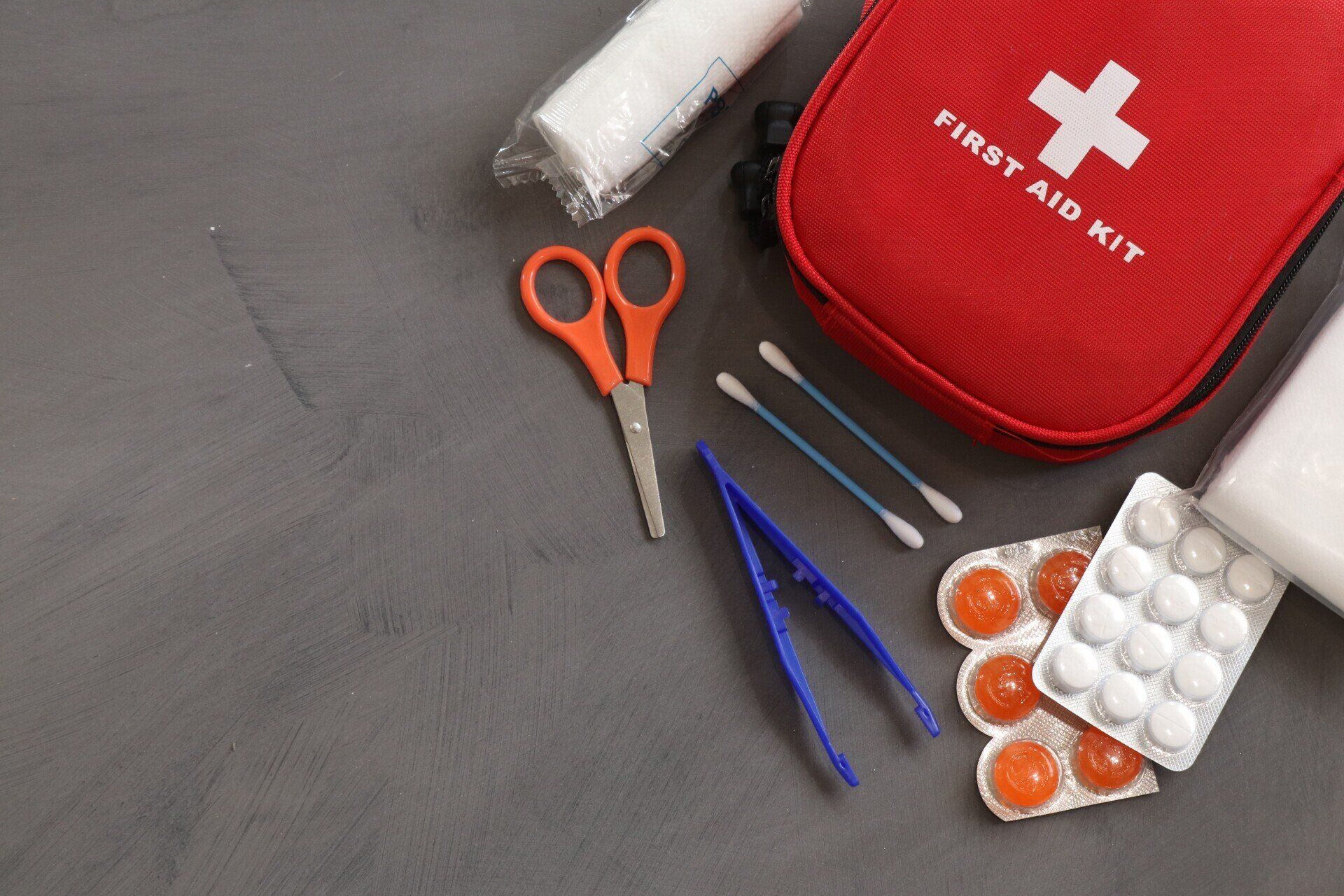 Rotes Erste-Hilfe-Set mit Schere, Pinzette, Wattestäbchen und Tablettenblisterpackungen auf grauer Oberfläche