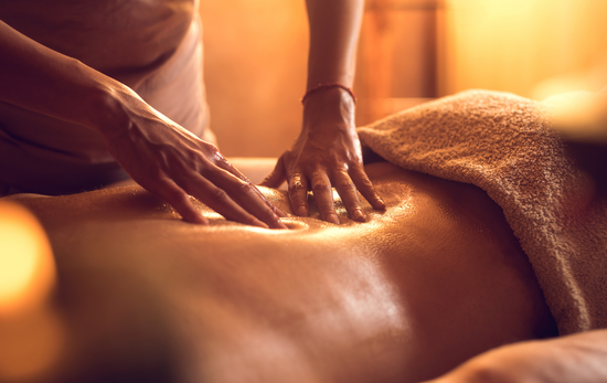 Hands massaging a person's back, covered by a towel, in a warmly lit spa setting.