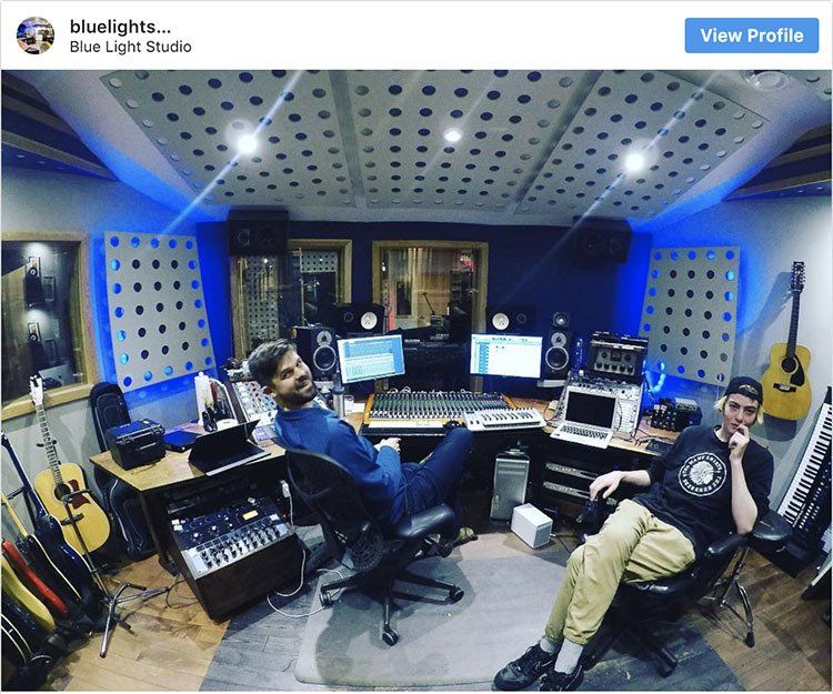 Two men relax in a recording studio with acoustic treatment. One sits in a chair, the other leans against the desk.