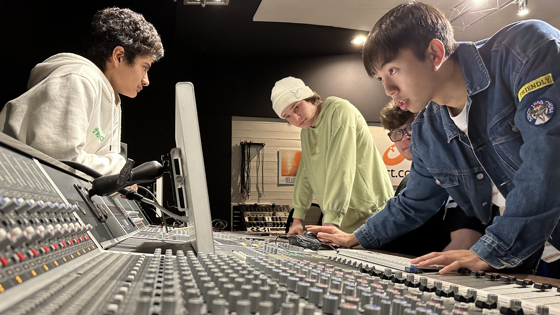 OIART students lean over a large audio mixing console in a studio, collaboratively adjusting knobs and sliders.