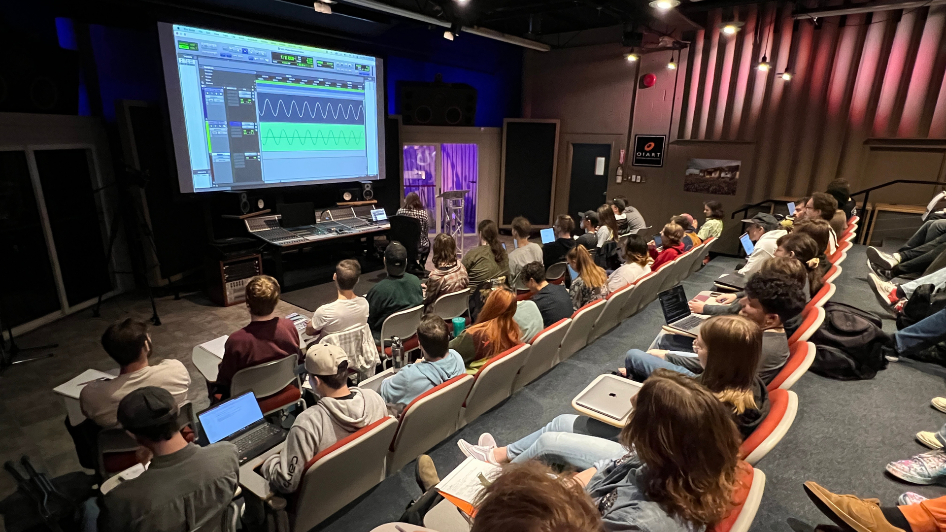 A classroom with students attending a music production lecture, observing a screen displaying music software.