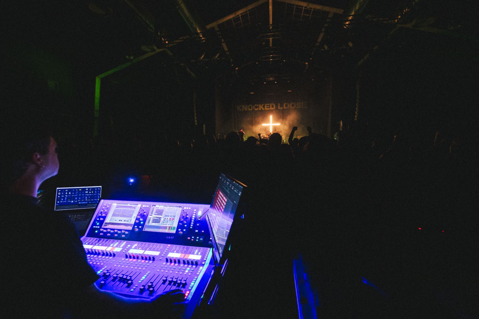 Dark concert venue with glowing blue sound equipment in foreground and bright stage lights in distance