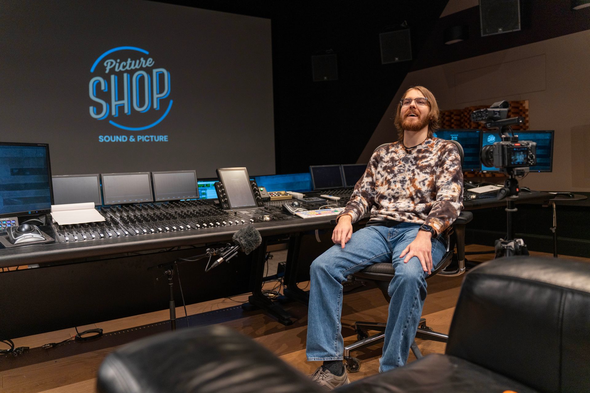 Gabriel Packer sitting in a studio with audio mixing consoles and monitors, under a “Podcast Shop” sign.
