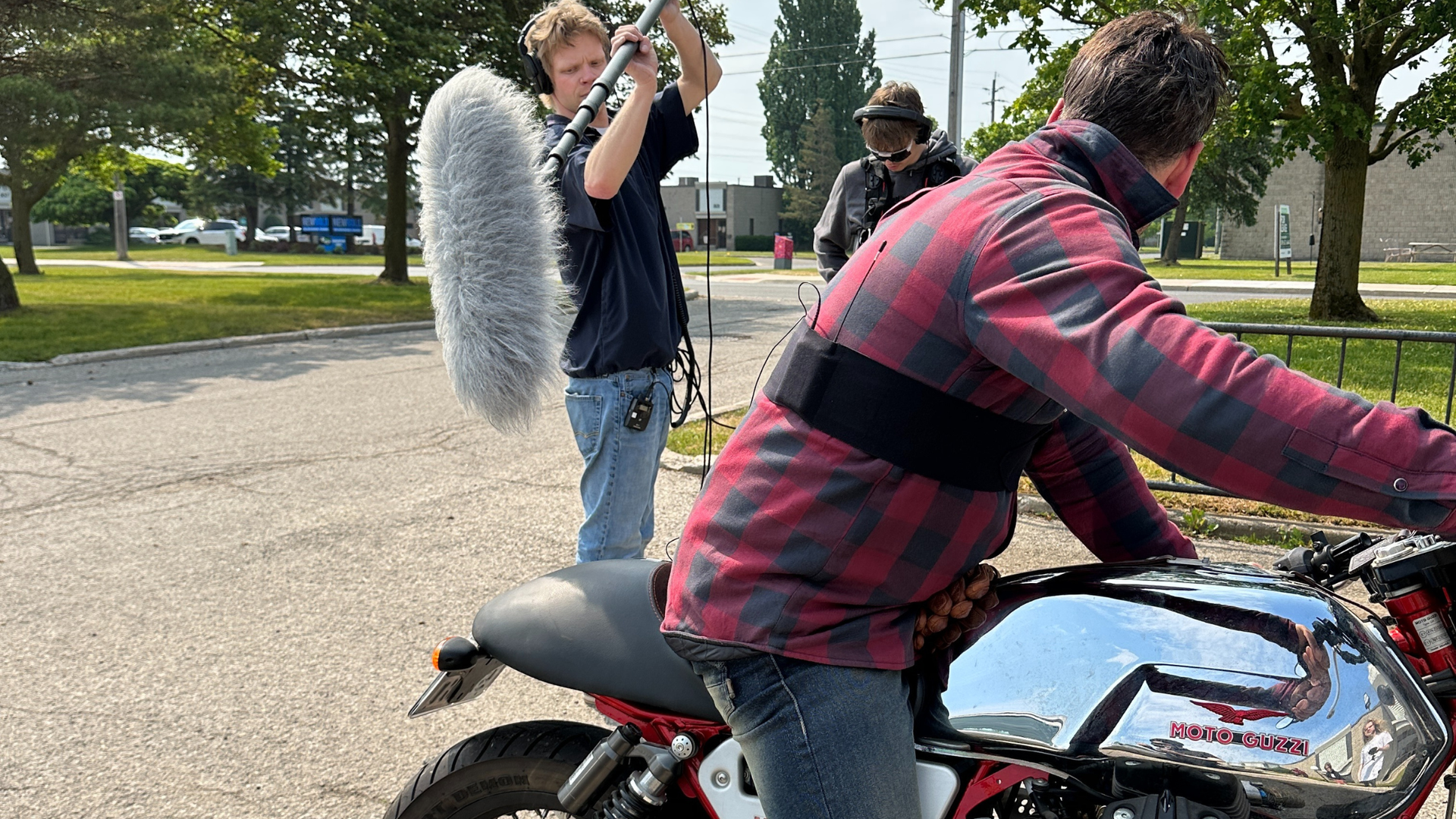 Filming a scene outdoors: Man on a motorcycle, sound operator with microphone, and another person in the background.
