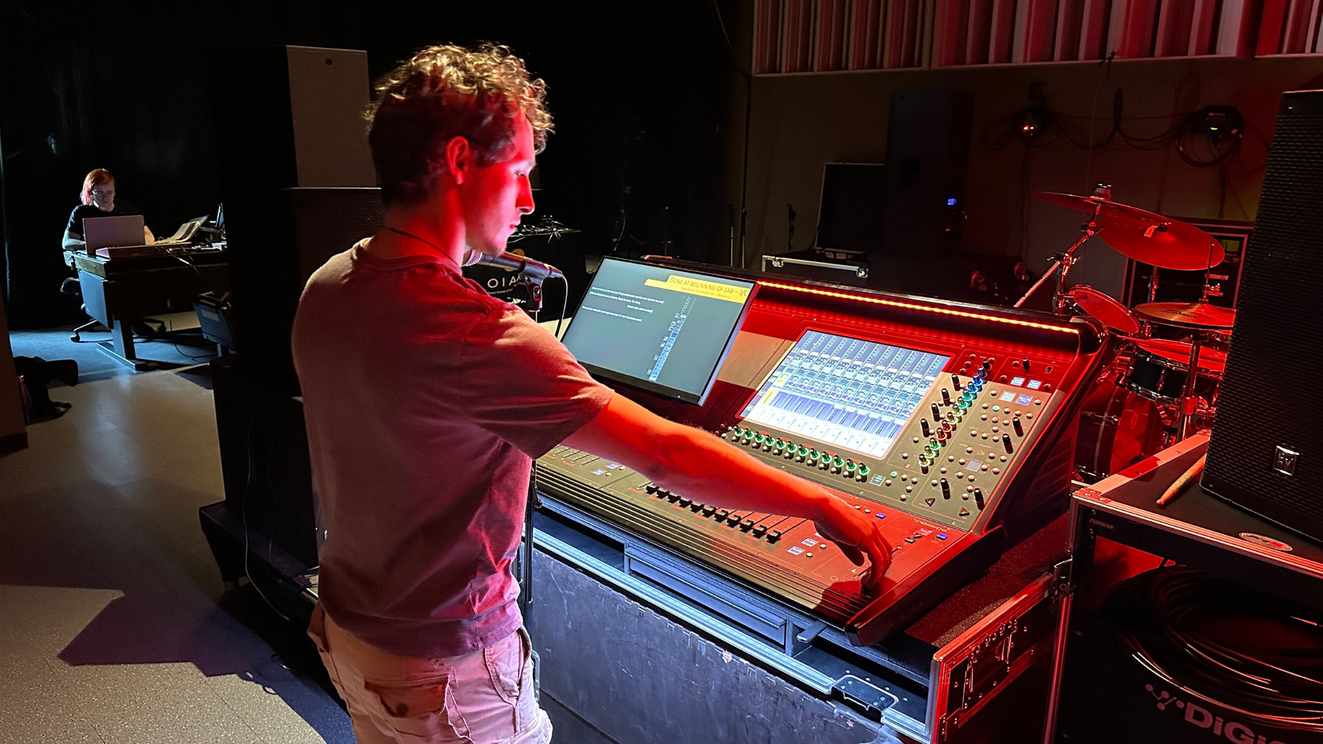 Person using a mixing console with lights. Another person in background. Inside a dim room.