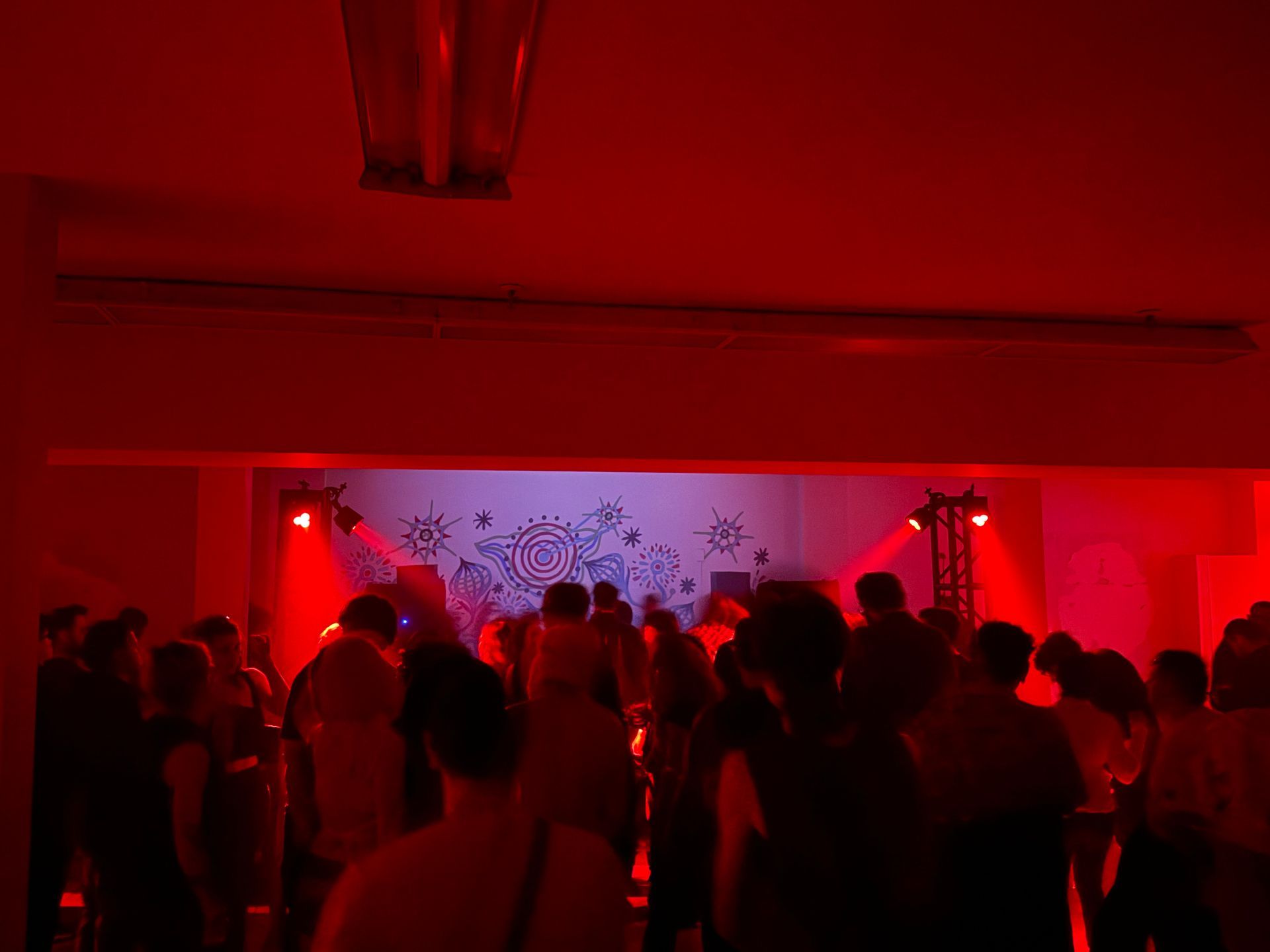 Crowded red-lit party room with people standing and dancing near a decorated wall