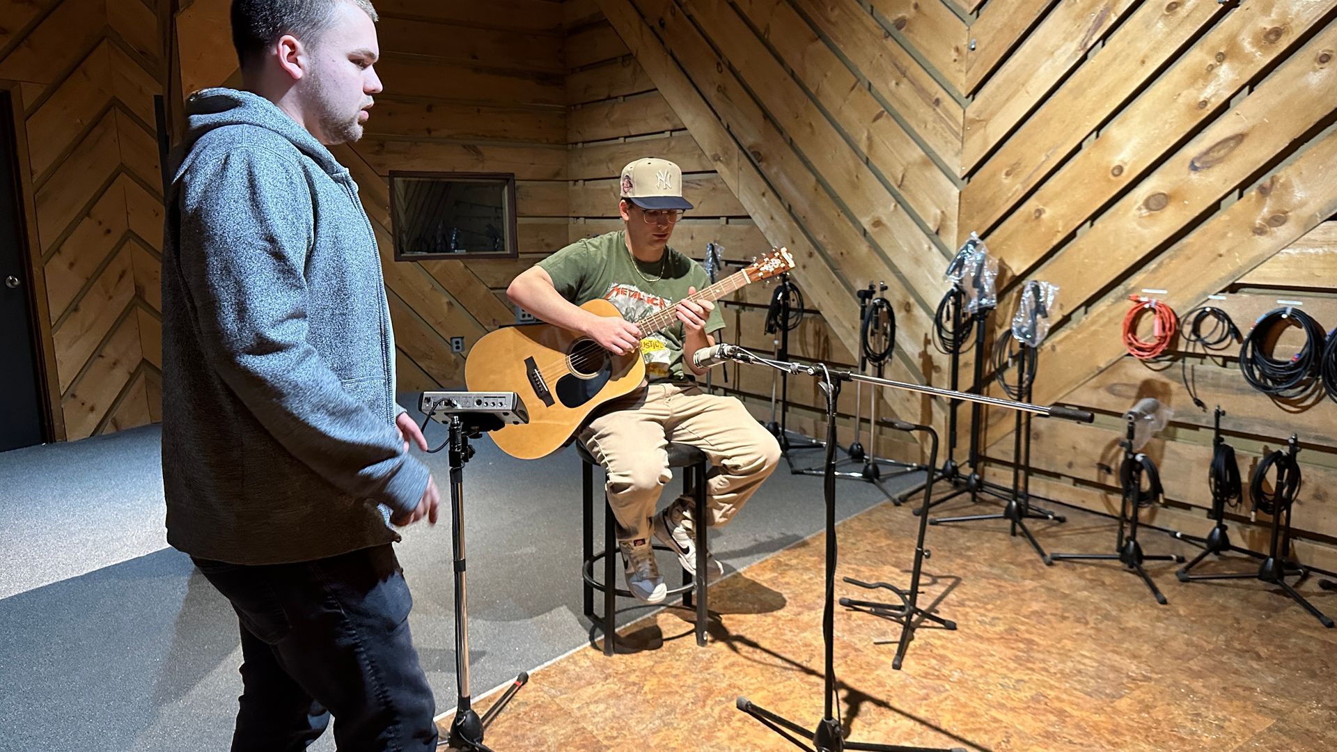 Avery playing acoustic guitar in a recording studio while another person watches nearby.