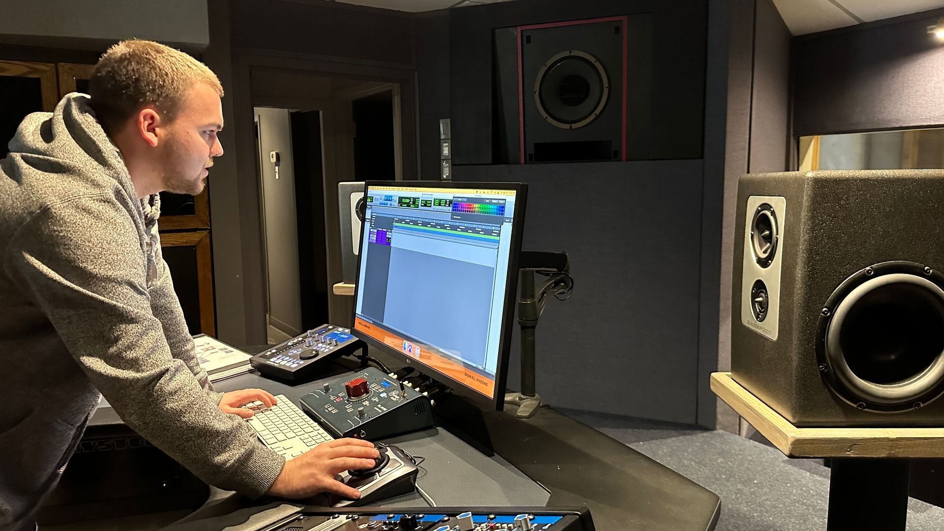 Avery Szumlanski editing audio in a studio, leaning over a mixing console beside a computer monitor and speakers.