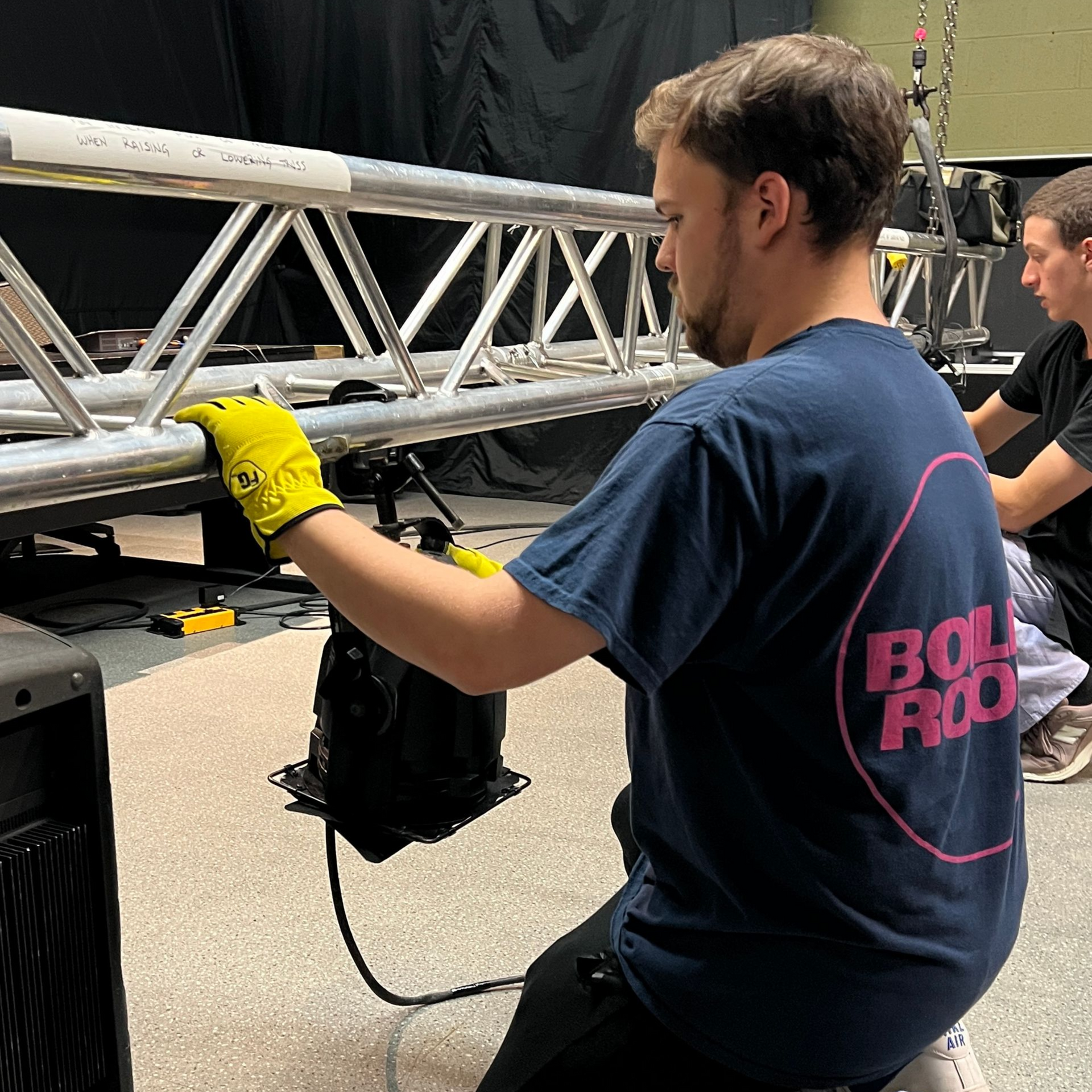 Avery a technician in yellow gloves adjusts a silver truss on stage during setup.