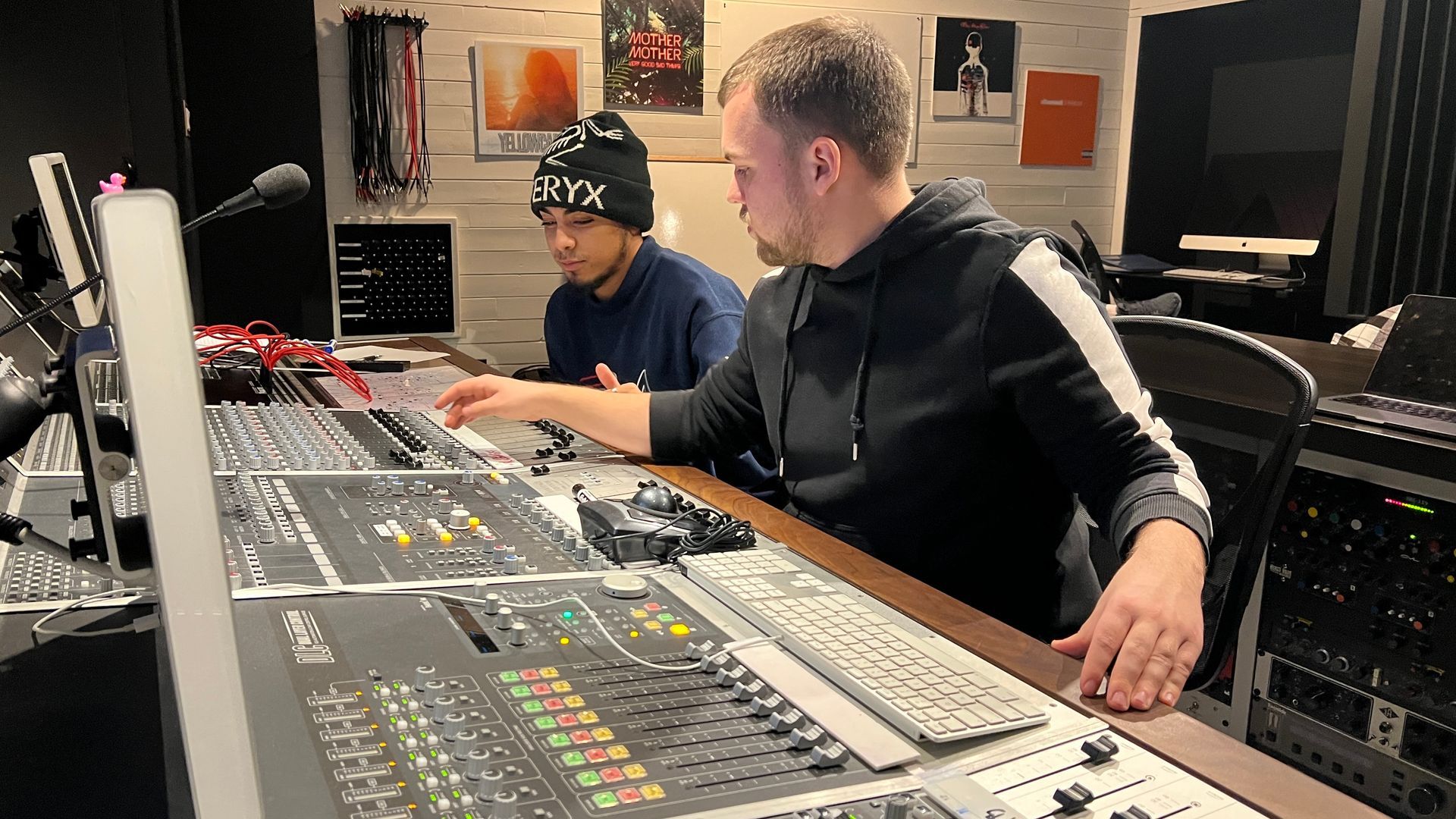 Avery & another working at a large audio mixing console in a studio, with monitors and equipment around them