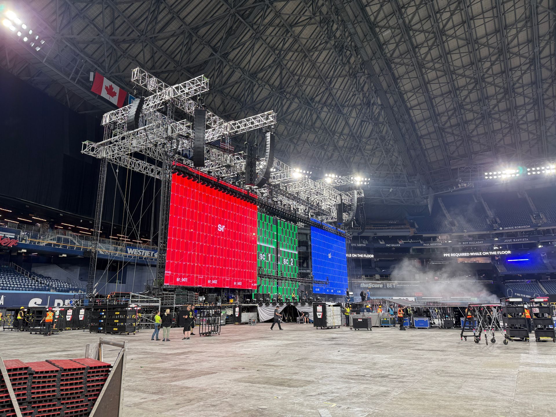 Large indoor stadium with a suspended stage, bright red, green, and blue LED screens, and crew on the floor