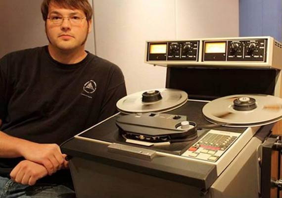 Douglas Beck in a black shirt is sitting in front of a tape machine
