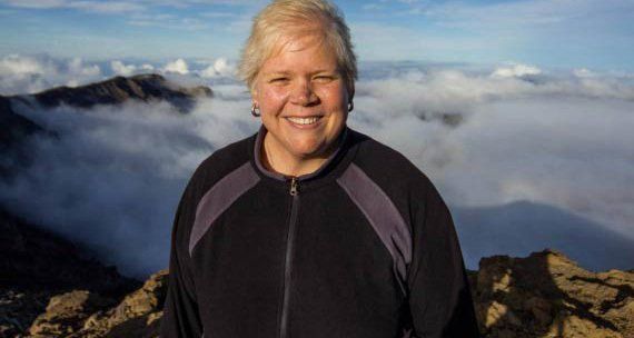 Jennifer Lewis is standing on top of a mountain with clouds in the background.