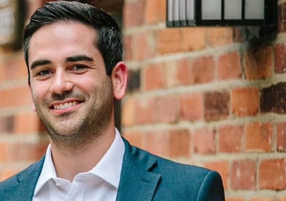 John Horwath in a suit and white shirt is smiling in front of a brick wall.