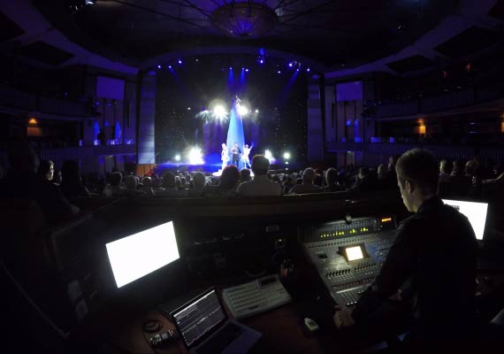 Josh Cuthbert at a soundboard in a theater, lights glowing the stage with performers & audience.