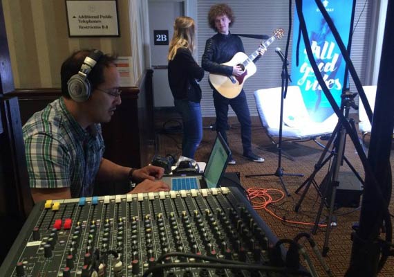 Keeron Tom wearing headphones sits in front of a mixer while a man plays a guitar