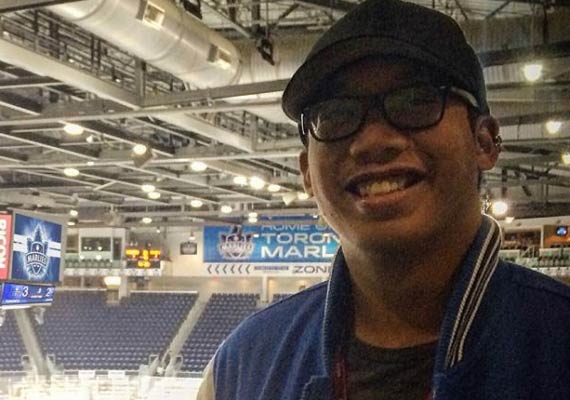Kelvin Tang smiling, in a hockey arena, blue jacket, background of seats and signage.