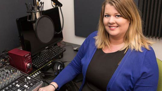 Paula Dineen in a blue sweater is sitting at a desk in front of a microphone.