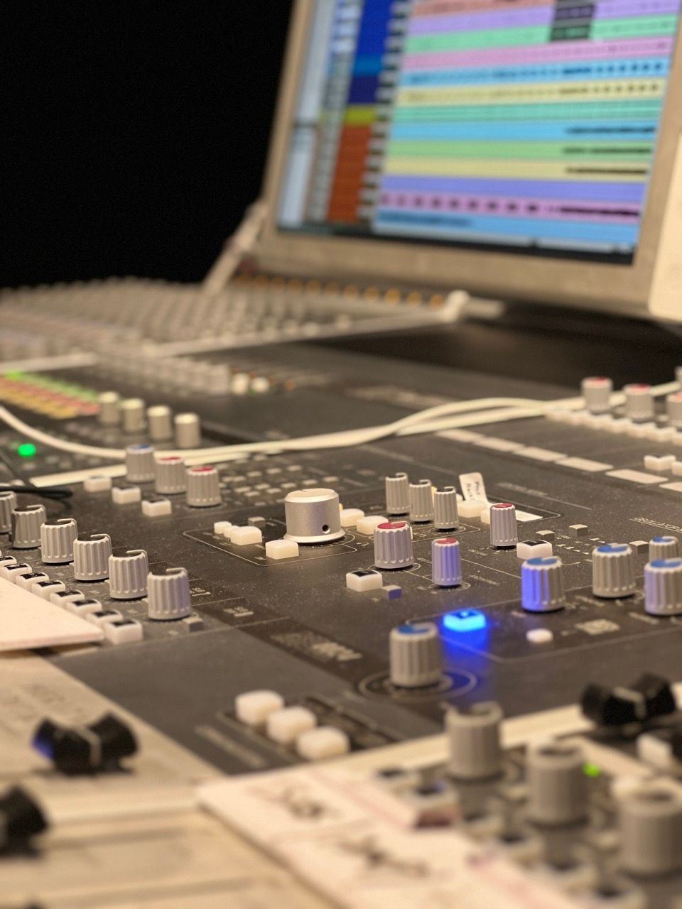 Close-up of a mixing console with knobs, faders, and a laptop displaying audio tracks in a recording studio.