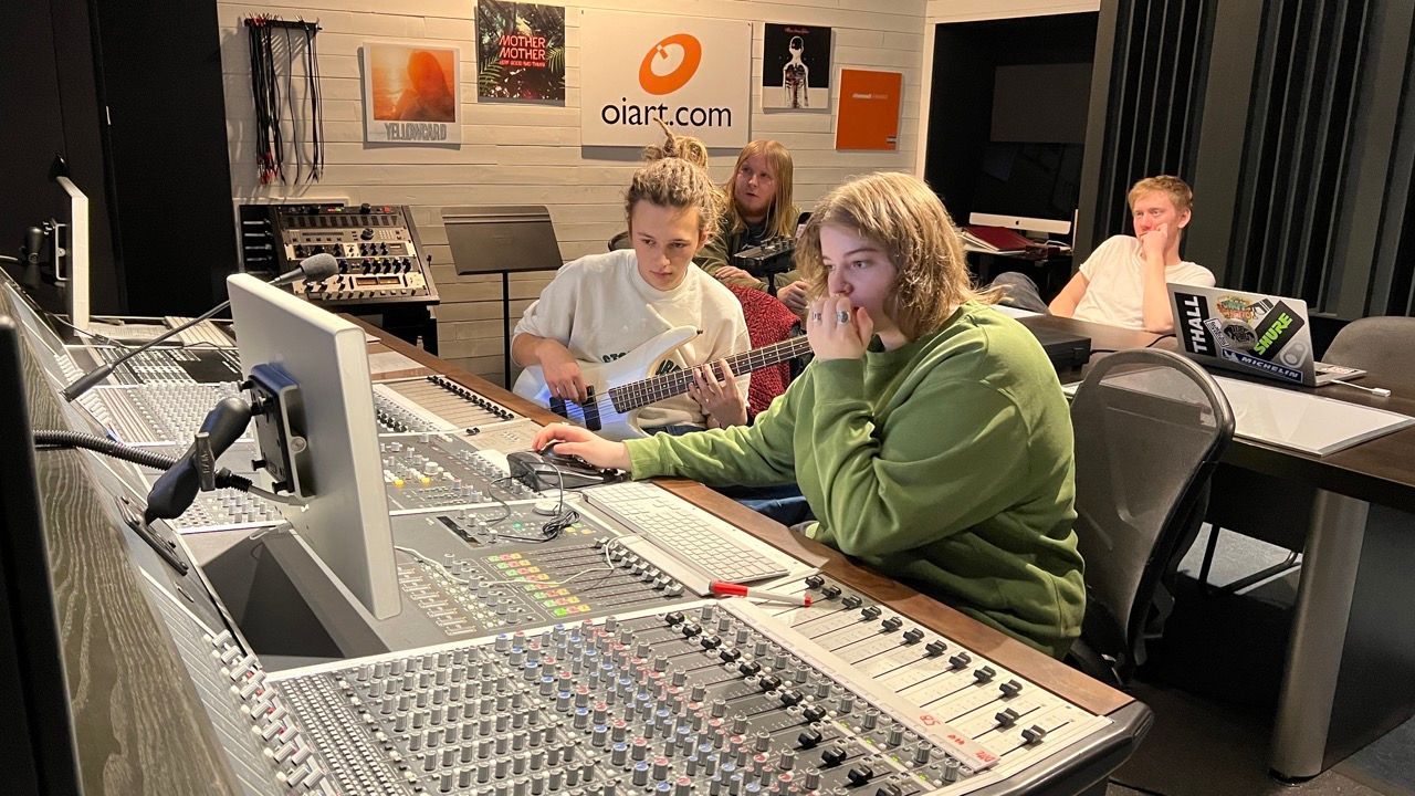 People in a recording studio. One plays guitar, another works at a mixing console, and others observe.