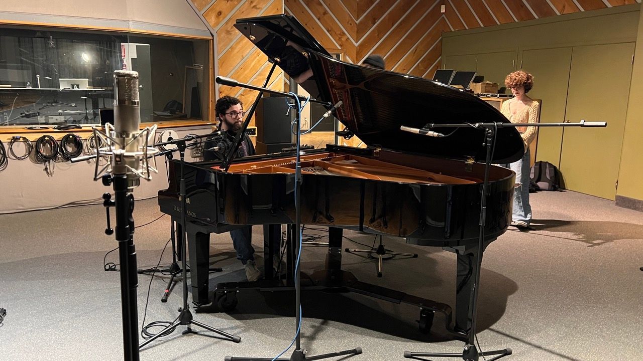 A person plays a grand piano in a recording studio, with microphones and a person standing nearby.