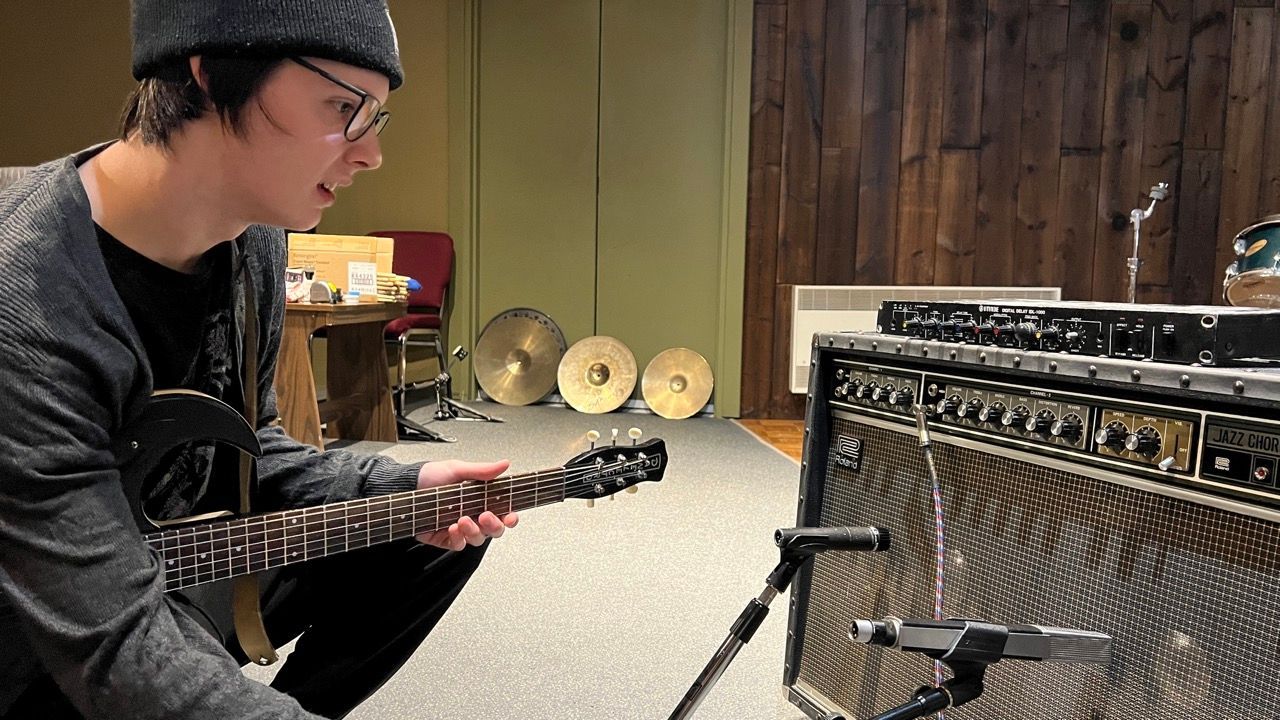 Musician plays guitar in studio, mic by amplifier. Room has wood paneling, cymbals, and equipment.